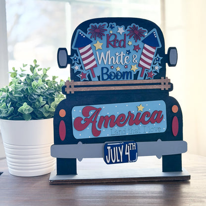 Decorative sign with 'Red White & Boom' and 'America Land that I Love' text, featuring American flags and July 4th elements.