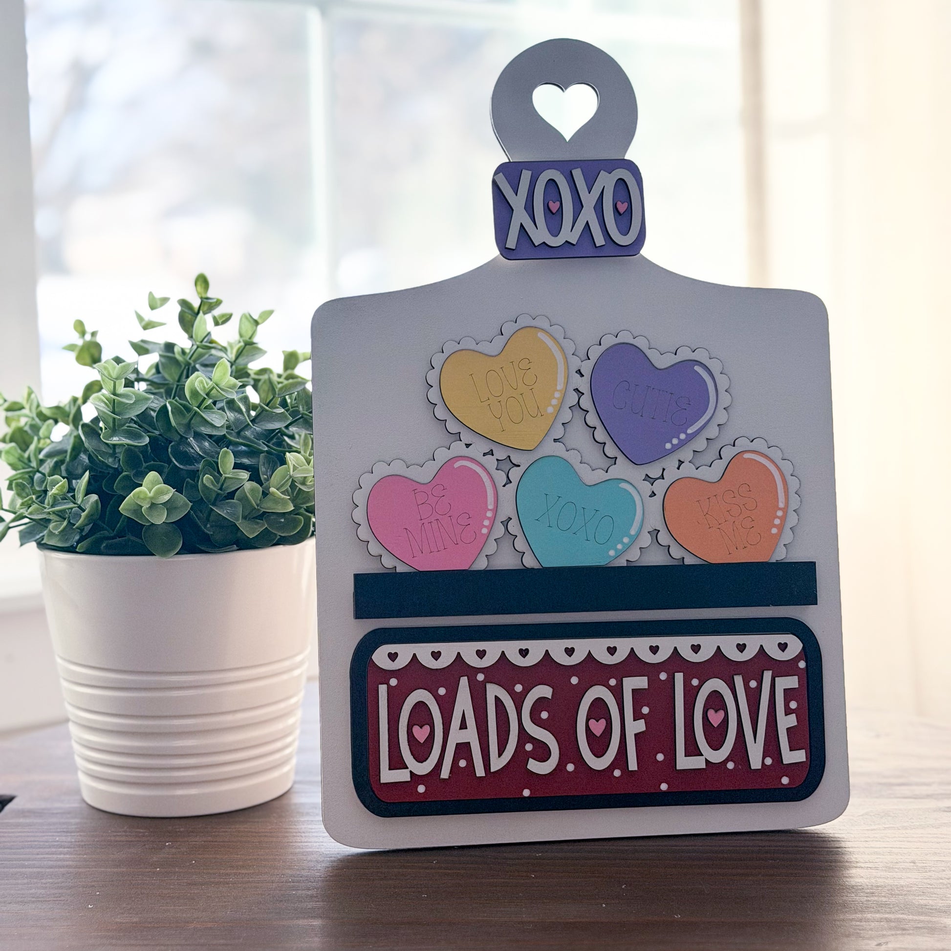 Decorative sign with colorful hearts and 'XOXO LOADS OF LOVE' text on a wooden surface with a plant in the background.