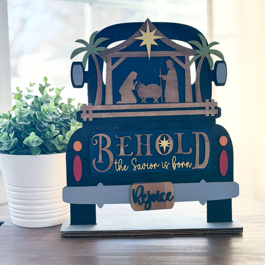 Decorative nativity scene in the shape of a truck with 'Behold the Savior is born' text, placed on a surface with a plant in the background.