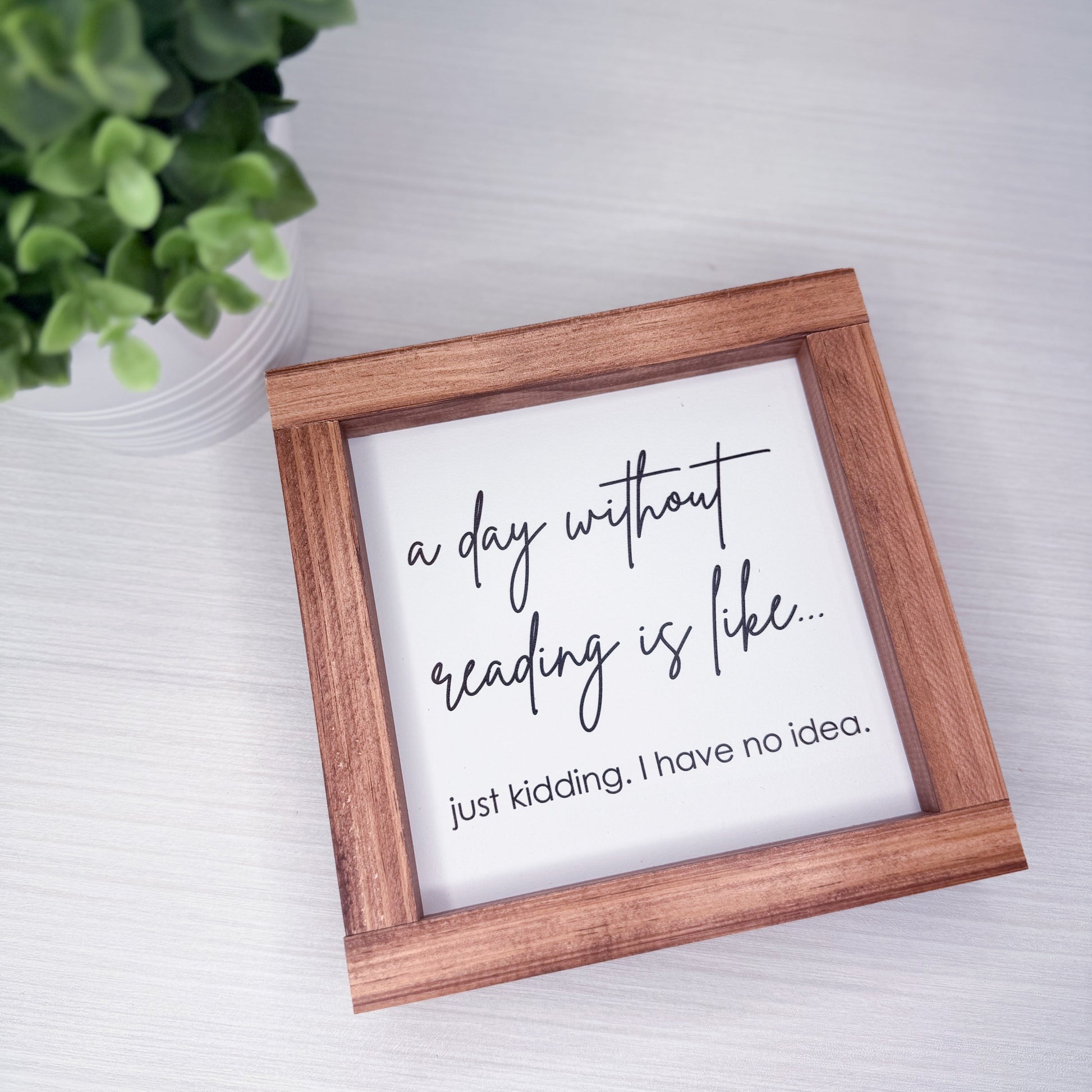Wooden-framed sign with humorous quote about reading on a light surface with a plant in the corner.