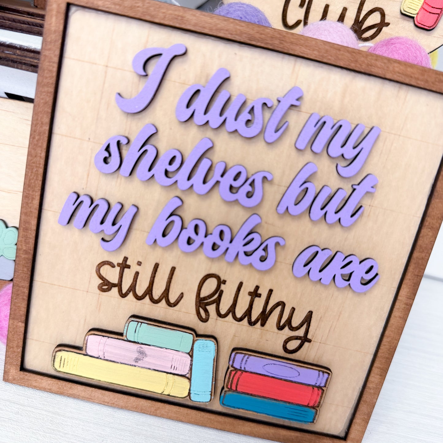 Wooden sign with colorful books and text 'I dust my shelves but my books are still filthy' on a wooden surface.