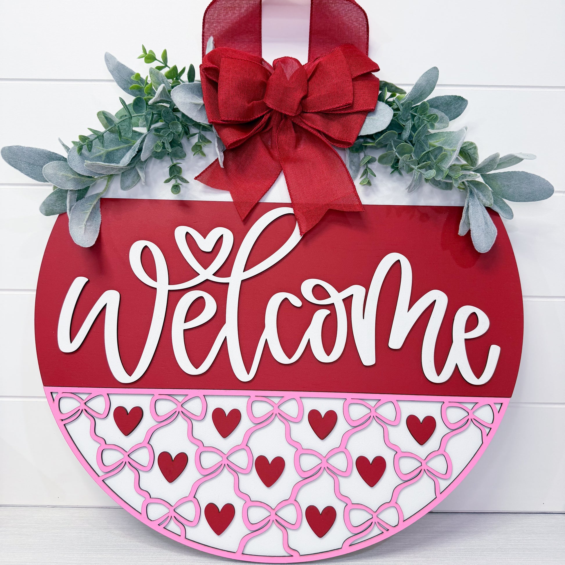 Decorative 'Welcome' sign with red bow, greenery, and heart patterns on a white background