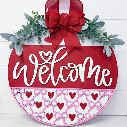 Decorative 'Welcome' sign with red bow, greenery, and heart patterns on a white background