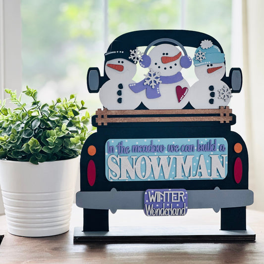 Decorative sign with snowmen in a car on a wooden surface with a plant in the background