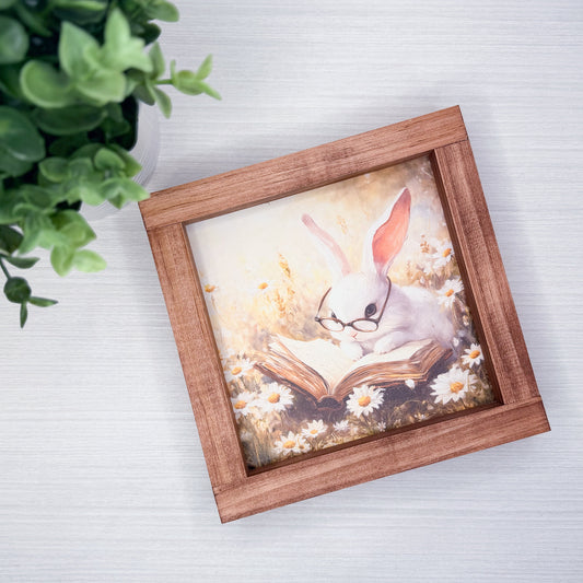 Framed artwork of a rabbit reading a book with flowers on a white surface.
