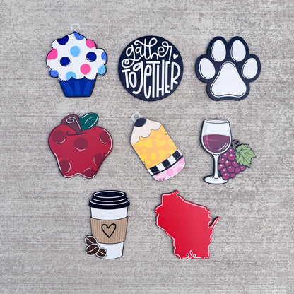 The pieces feature various designs including a blue and pink polka-dotted cupcake, a black circular pin reading 'gather together' in white script, a white paw print, a red apple, a yellow and pink striped ice cream cone, a wine glass with purple grapes, a brown coffee cup, and a red outline of Wisconsin state.