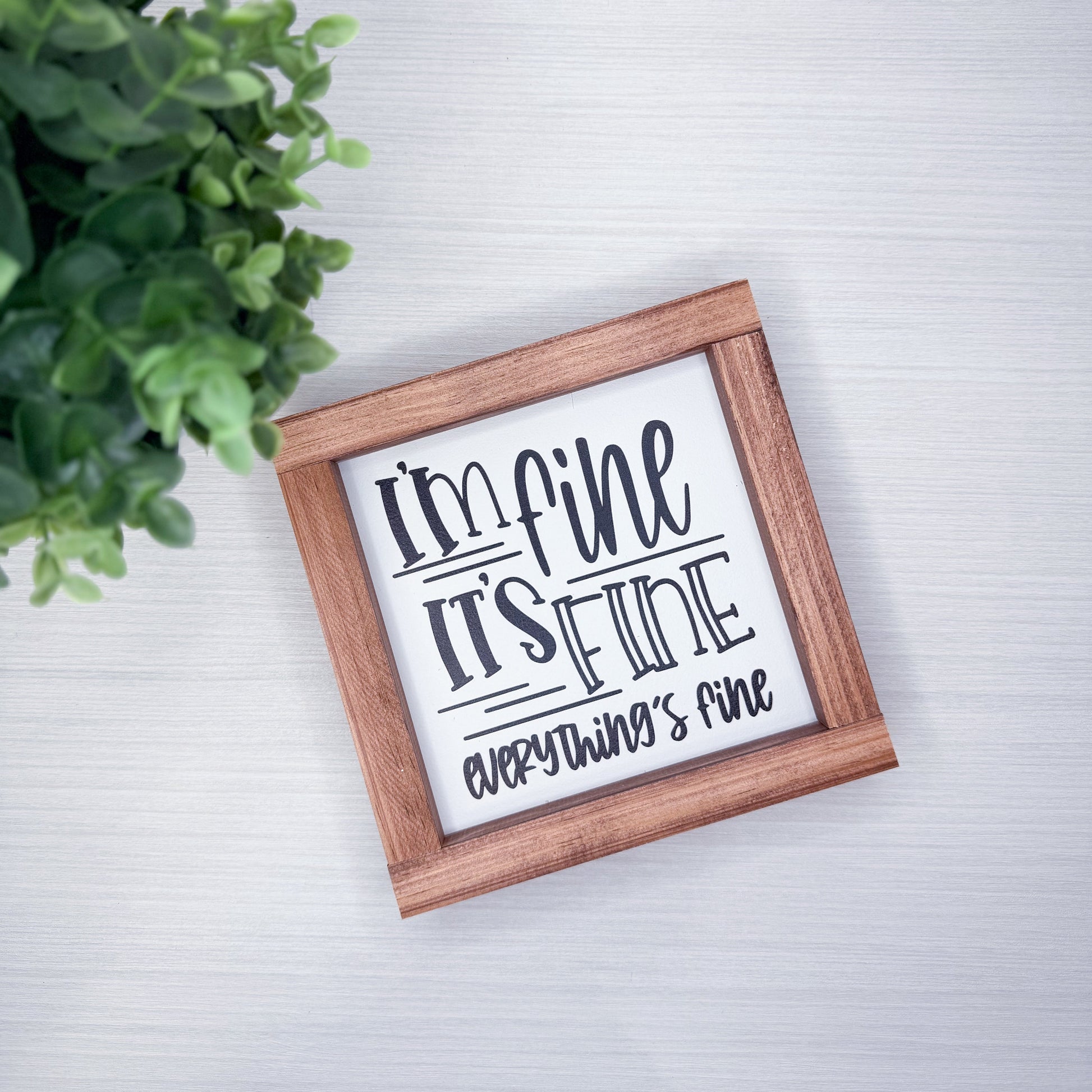 Wooden-framed sign with text 'I'm fine, it's fine, everything is fine' on a light background with a plant.