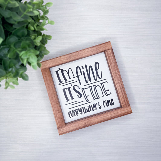 Wooden-framed sign with text 'I'm fine, it's fine, everything is fine' on a light background with a plant.
