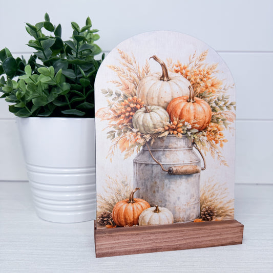 Arched wooden decorative sign with a fall harvest design featuring pumpkins, wheat, and a galvanized milk canister on a wooden base, styled next to a potted green plant.