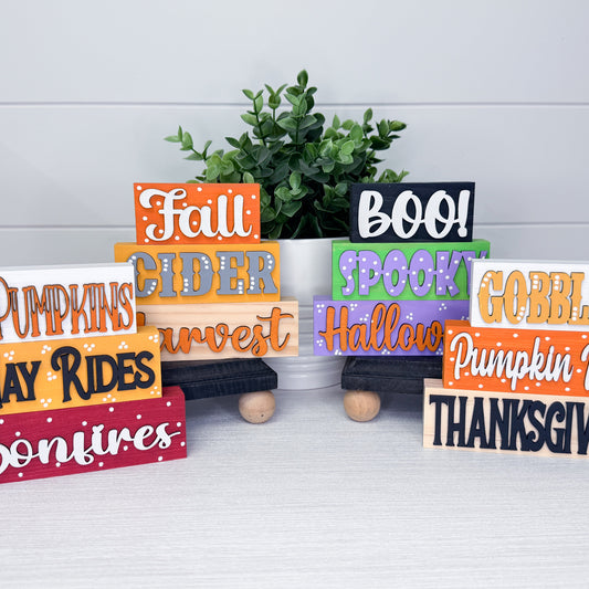 A set of colorful wooden decorative signs with autumn and fall-themed words such as 'Fall,' 'BOO!,' 'Spooky,' 'Halloween,' 'Pumpkins,' 'Cider,' 'Harvest,' 'Gobble,' 'Thanksgiving,' and 'Moonfires,' arranged on small black and wooden carts in front of a white plant pot with green foliage.