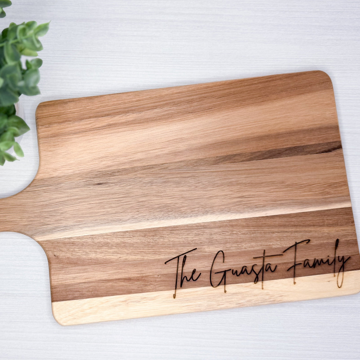 Wooden cutting board with 'The Guasta Family' engraving on a white surface with a green plant.