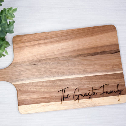 Wooden cutting board with 'The Guasta Family' engraving on a white surface with a green plant.