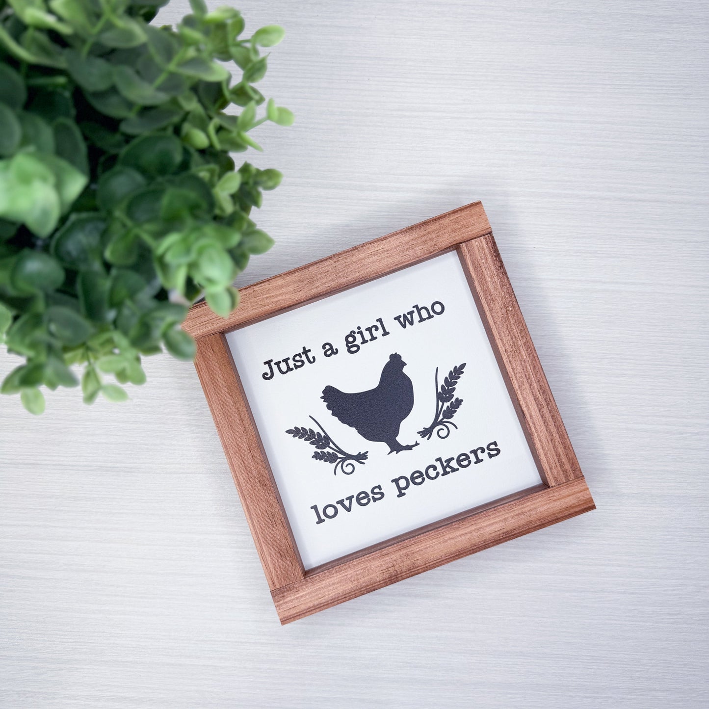 Small wooden-framed sign with a chicken illustration and text on a white surface with greenery.