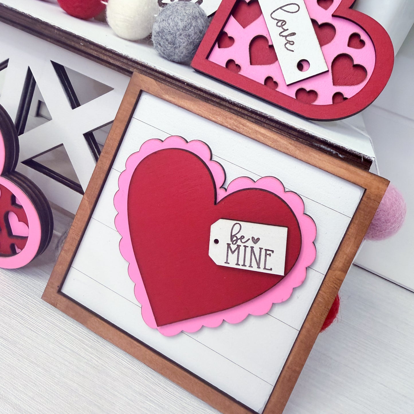 Heart-shaped card with 'be mine' tag on a wooden frame, surrounded by decorative items.