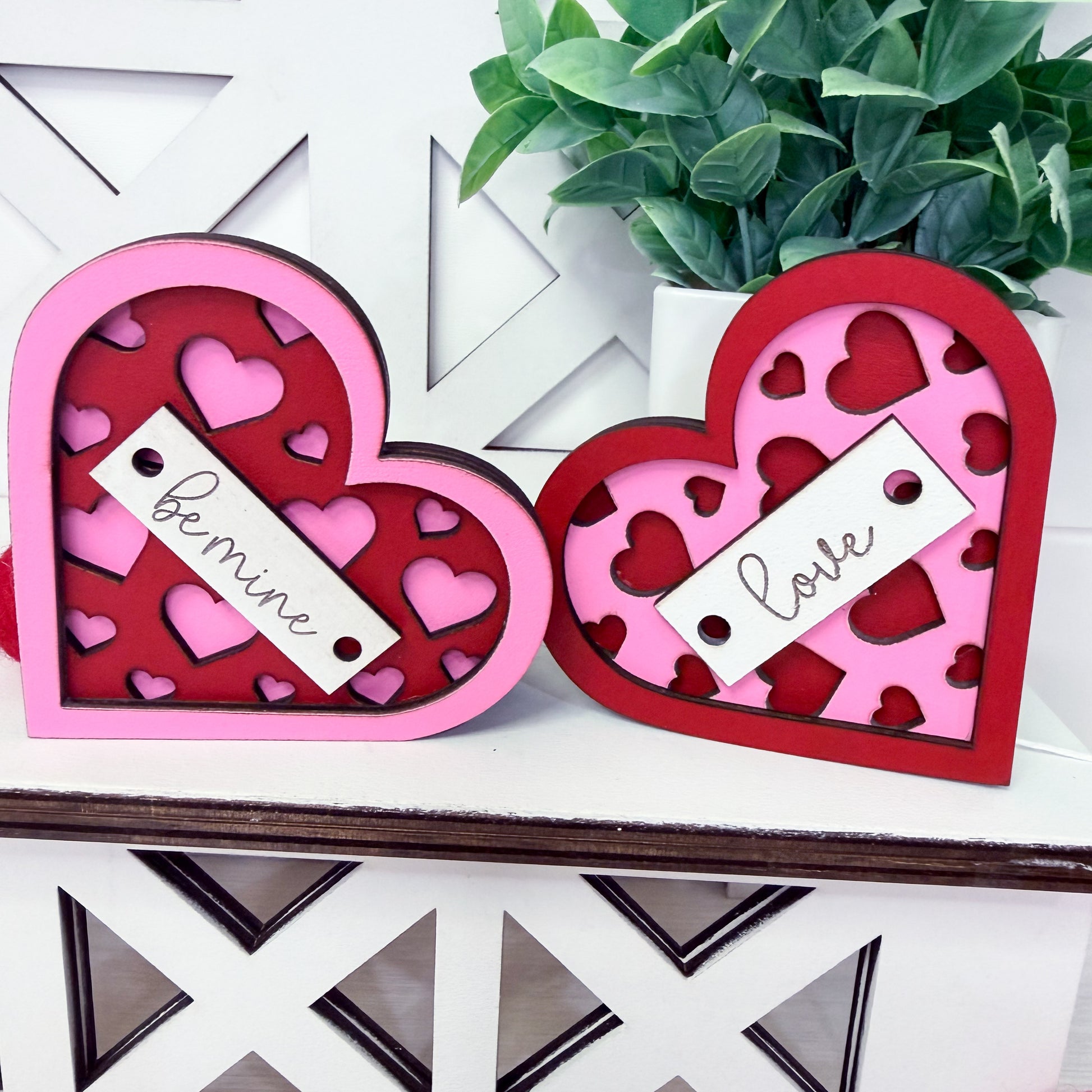 Two heart-shaped decorative boxes with 'be mine' and 'love' tags on a white surface with a plant in the background.