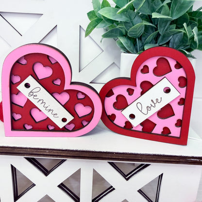 Two heart-shaped decorative boxes with 'be mine' and 'love' tags on a white surface with a plant in the background.