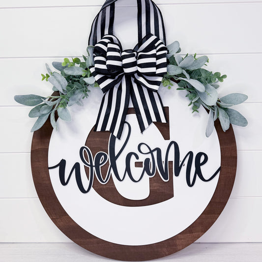 Decorative wreath with 'welcome' sign, black and white striped bow, and greenery on a white background