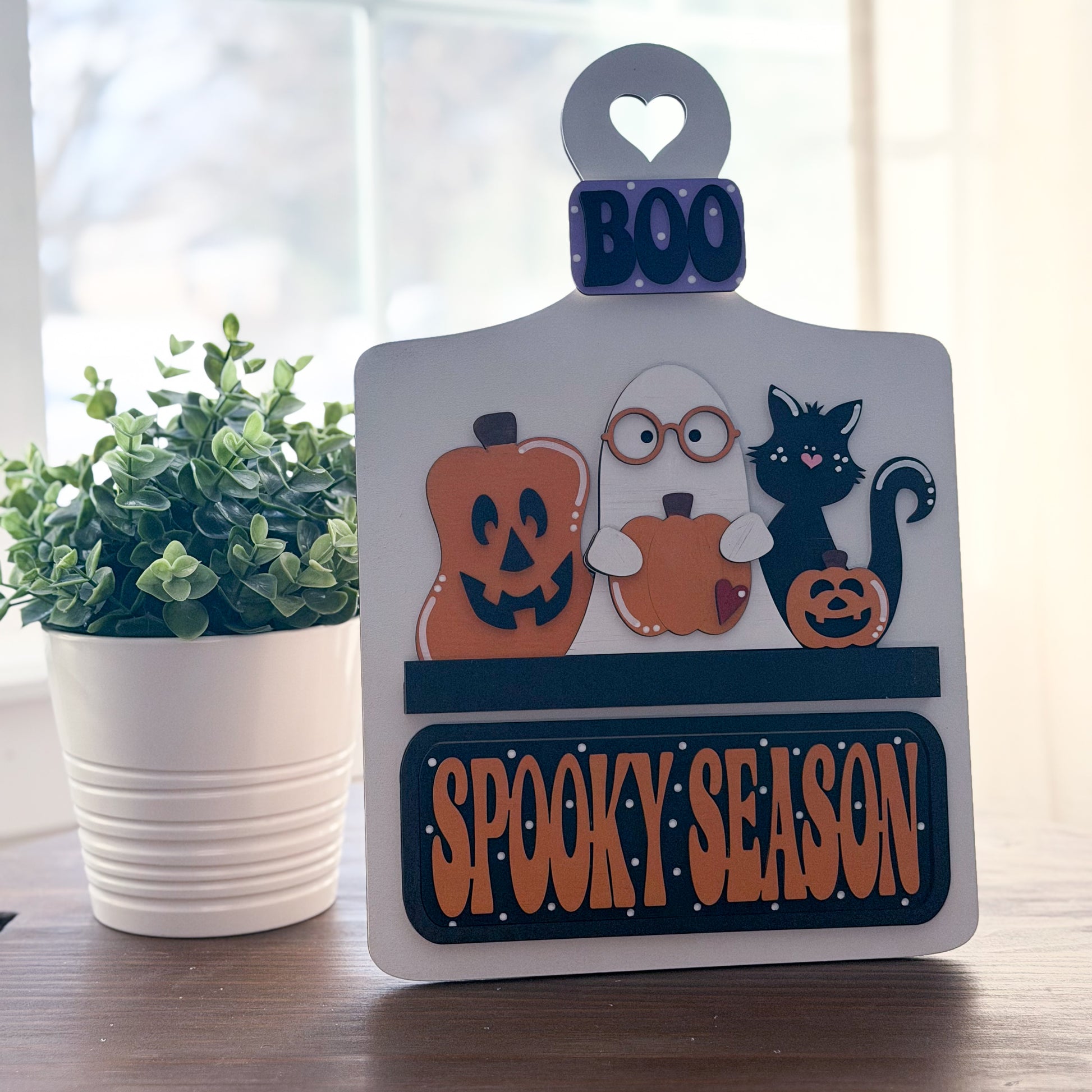 Halloween-themed decorative sign with pumpkins, ghost, and cat on a wooden surface with a plant in the background.