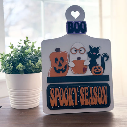 Halloween-themed decorative sign with pumpkins, ghost, and cat on a wooden surface with a plant in the background.