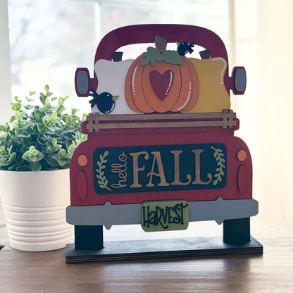 Decorative sign shaped like a red truck with pumpkins and 'hello Fall' text on a wooden surface.