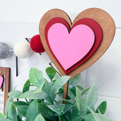 Heart-shaped wooden stake with pink center and brown outer rim, placed among green plants.