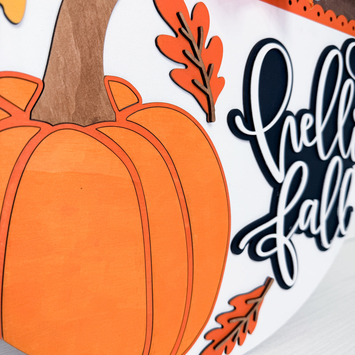 Close-up of a fall-themed decorative sign featuring a large orange pumpkin, orange leaves, and white script text reading 'hello fall' on a white background.