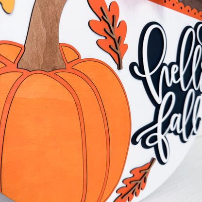 Close-up of a fall-themed decorative sign featuring a large orange pumpkin, orange leaves, and white script text reading 'hello fall' on a white background.