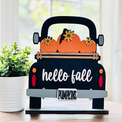 A decorative wood truck with 'hello fall' and 'pumpkin' text, carrying pumpkins on its bed, placed on a table.