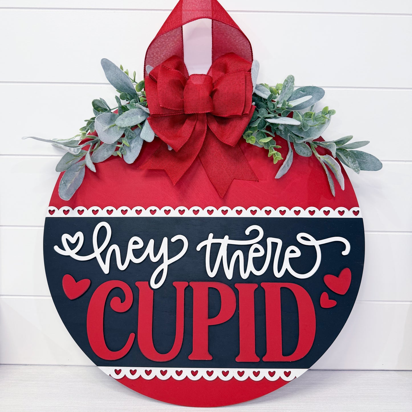 Decorative sign with 'Hey there Cupid' text on a red and black background with greenery and a bow.
