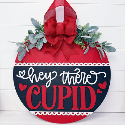 Decorative sign with 'Hey there Cupid' text on a red and black background with greenery and a bow.