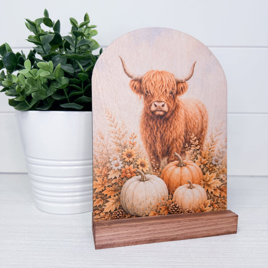 A decorative arched wooden plaque featuring a painted Highland cow standing among pumpkins and autumn foliage, displayed on a small wooden base next to a potted green plant.