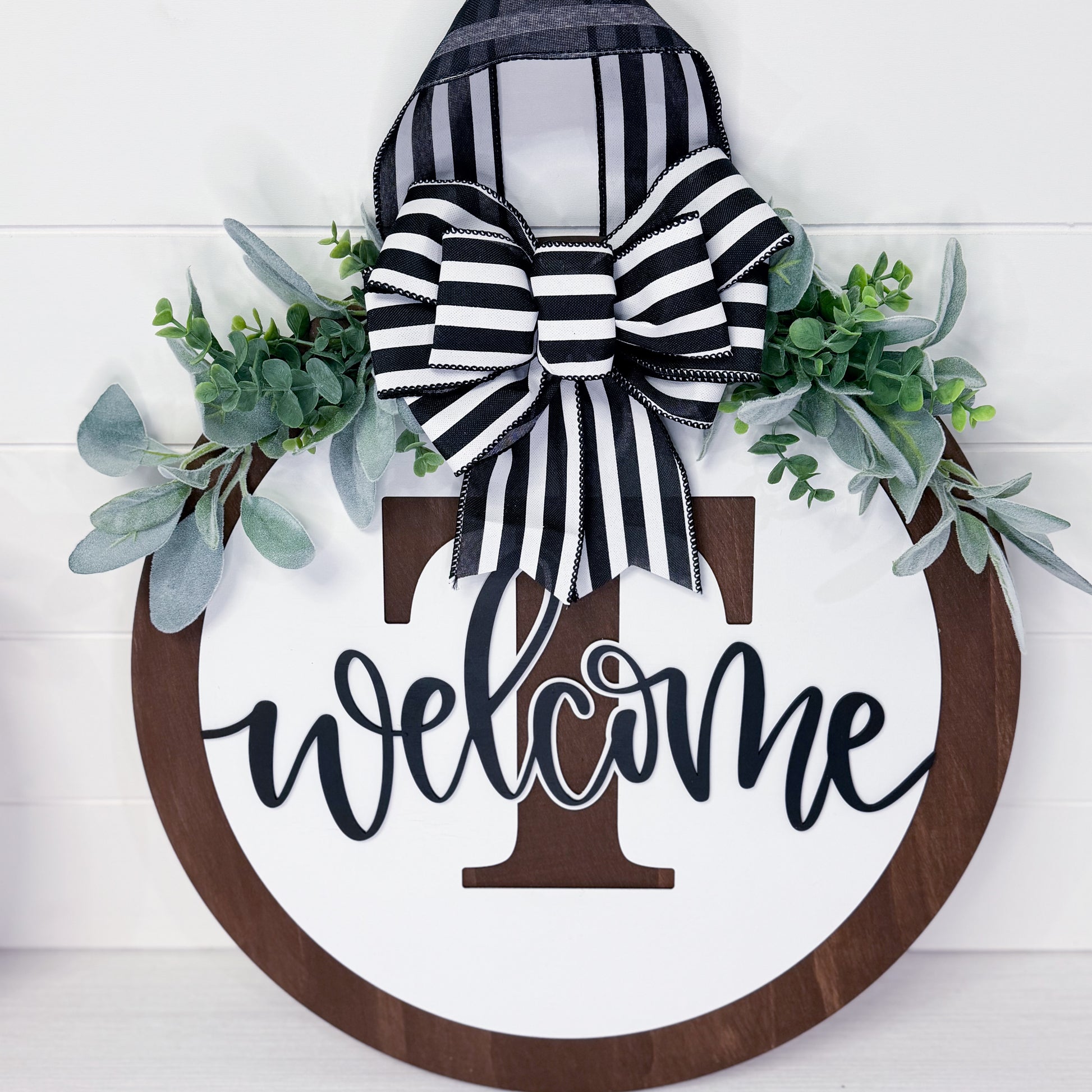 Decorative wreath with 'welcome' sign, striped bow, and greenery on a white background