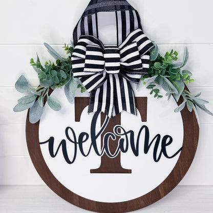 Decorative wreath with 'welcome' sign, striped bow, and greenery on a white background