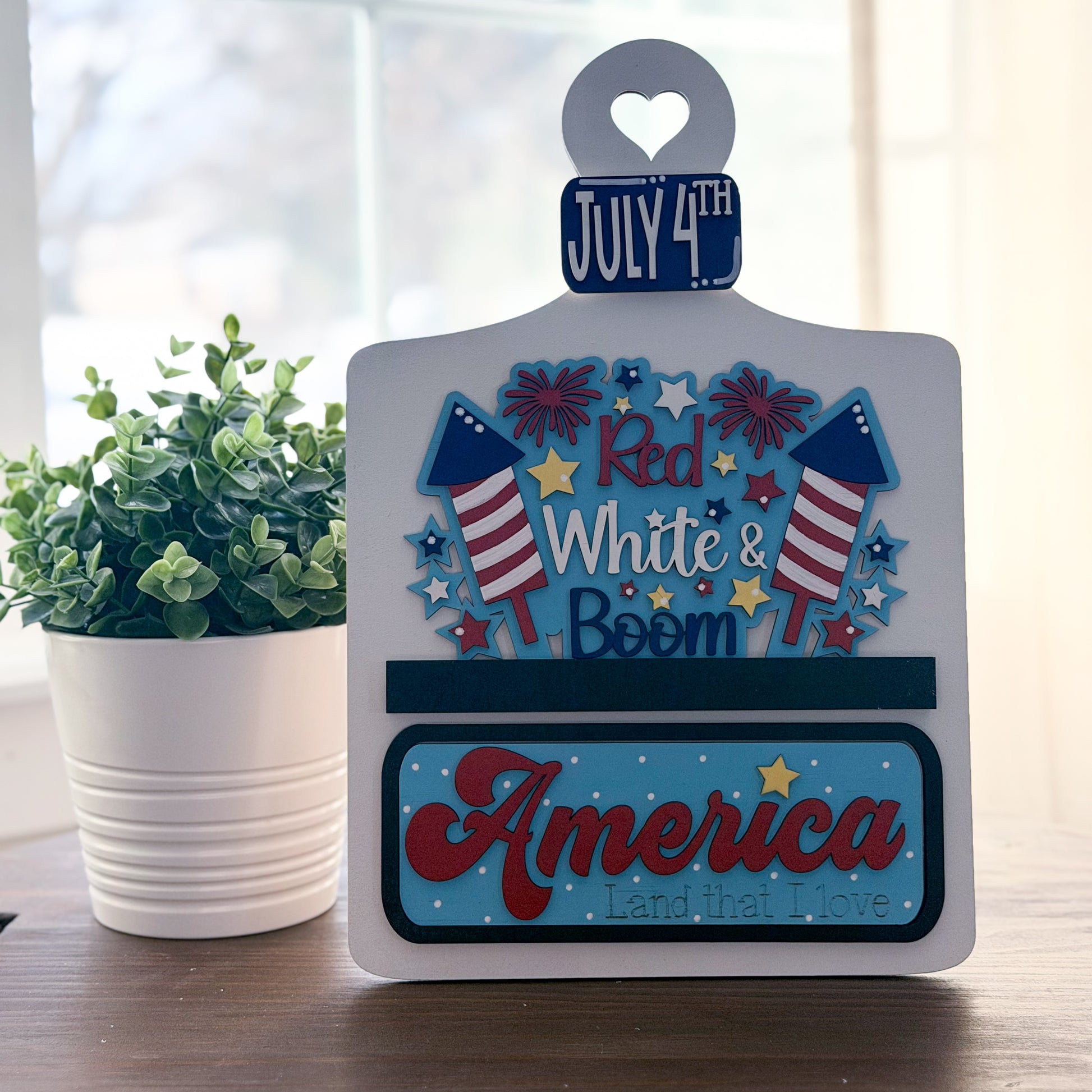 Decorative sign with July 4th theme on a table with a plant in the background