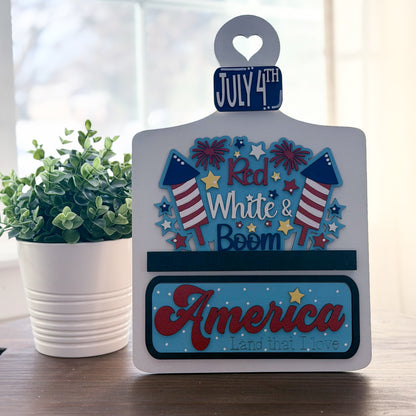 Decorative sign with July 4th theme on a table with a plant in the background