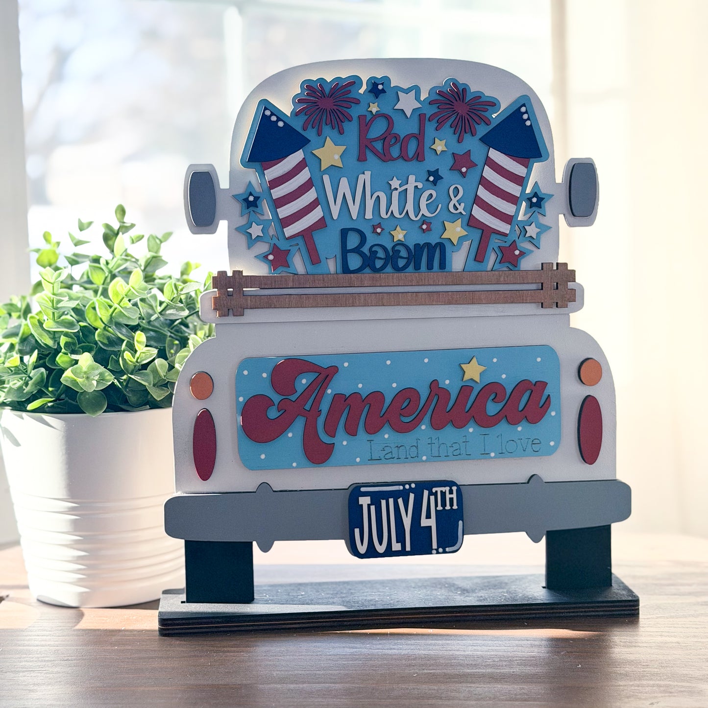 Decorative sign with 'Red White & Boom' and 'America Land that I Love' text, featuring American flags and July 4th theme.
