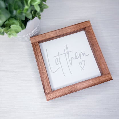 Wooden frame with 'Let them' text on a white surface with a plant in the corner.