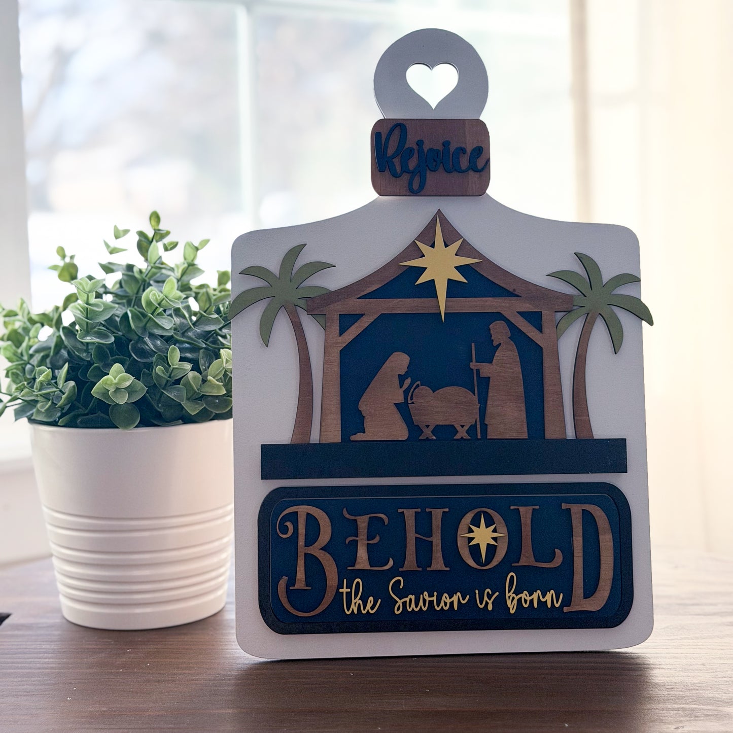 Decorative Christmas sign with nativity scene and 'Behold the Saviour is born' text, placed on a surface with a plant in the background.