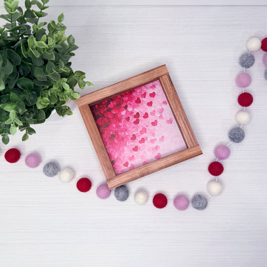 Wooden frame with pink heart-patterned paper, green plant, and colorful pom-pom garland on a white surface
