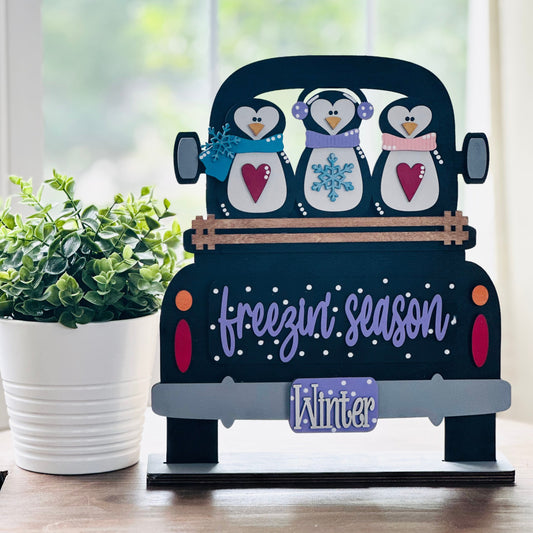 Decorative sign with penguins in a truck on a wooden surface with a plant in the background