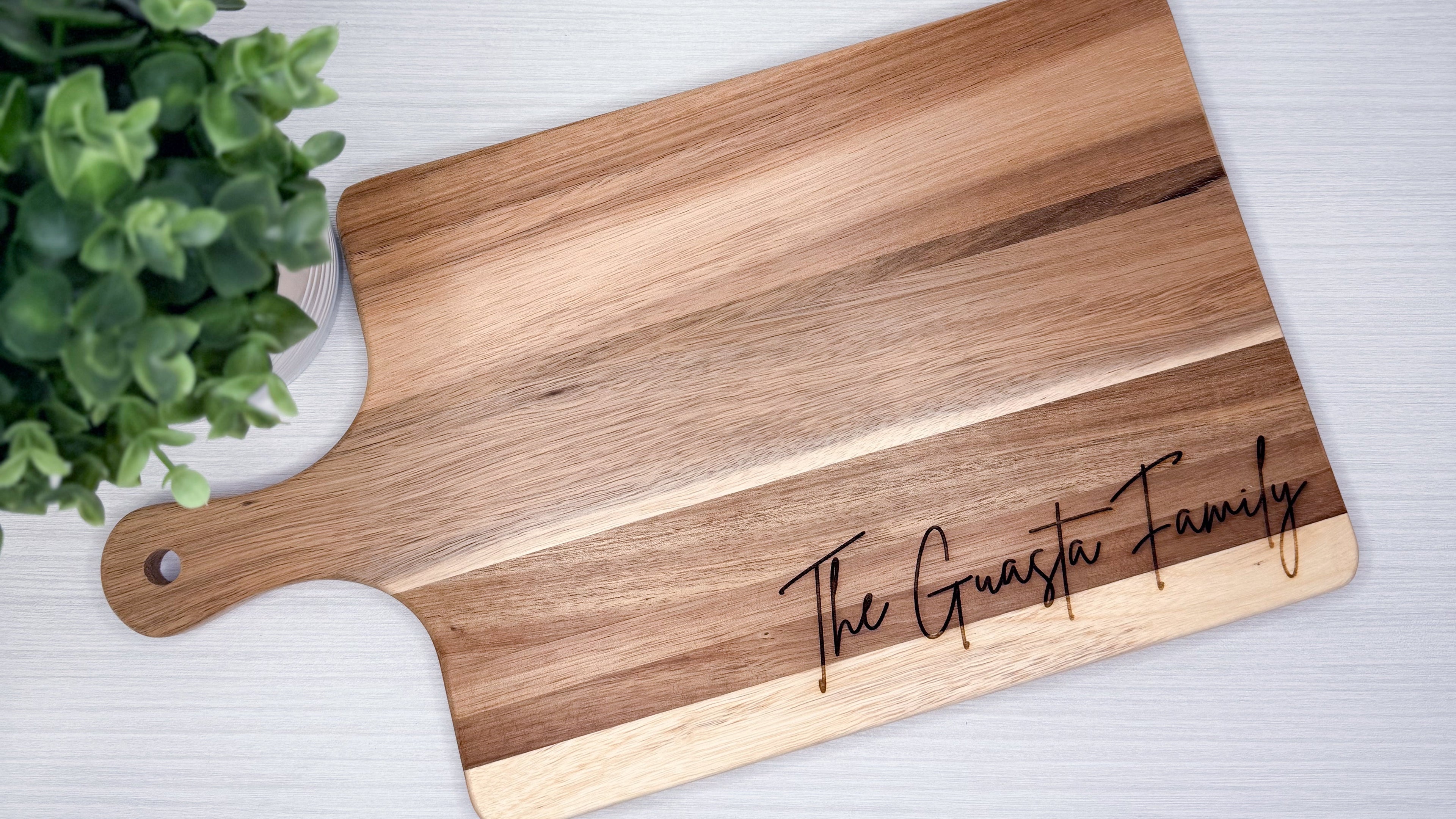 Wooden cutting board with 'The Guasta Family' engraving on a light gray surface with a plant in the corner.