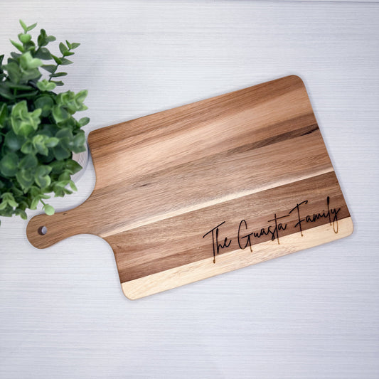 Wooden cutting board with 'The Guasta Family' engraving on a light gray surface with a plant in the corner.