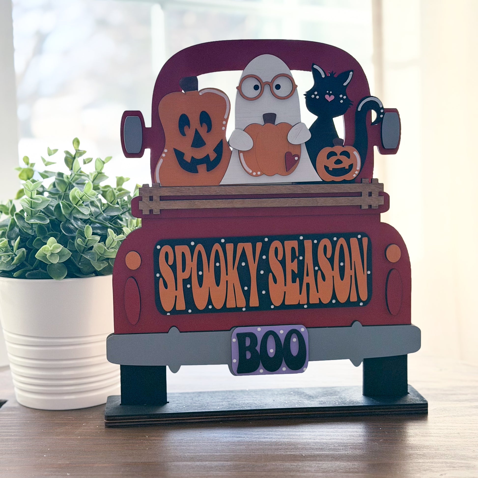 Halloween-themed decorative sign with a truck, ghost, cat, and pumpkin on a wooden surface.