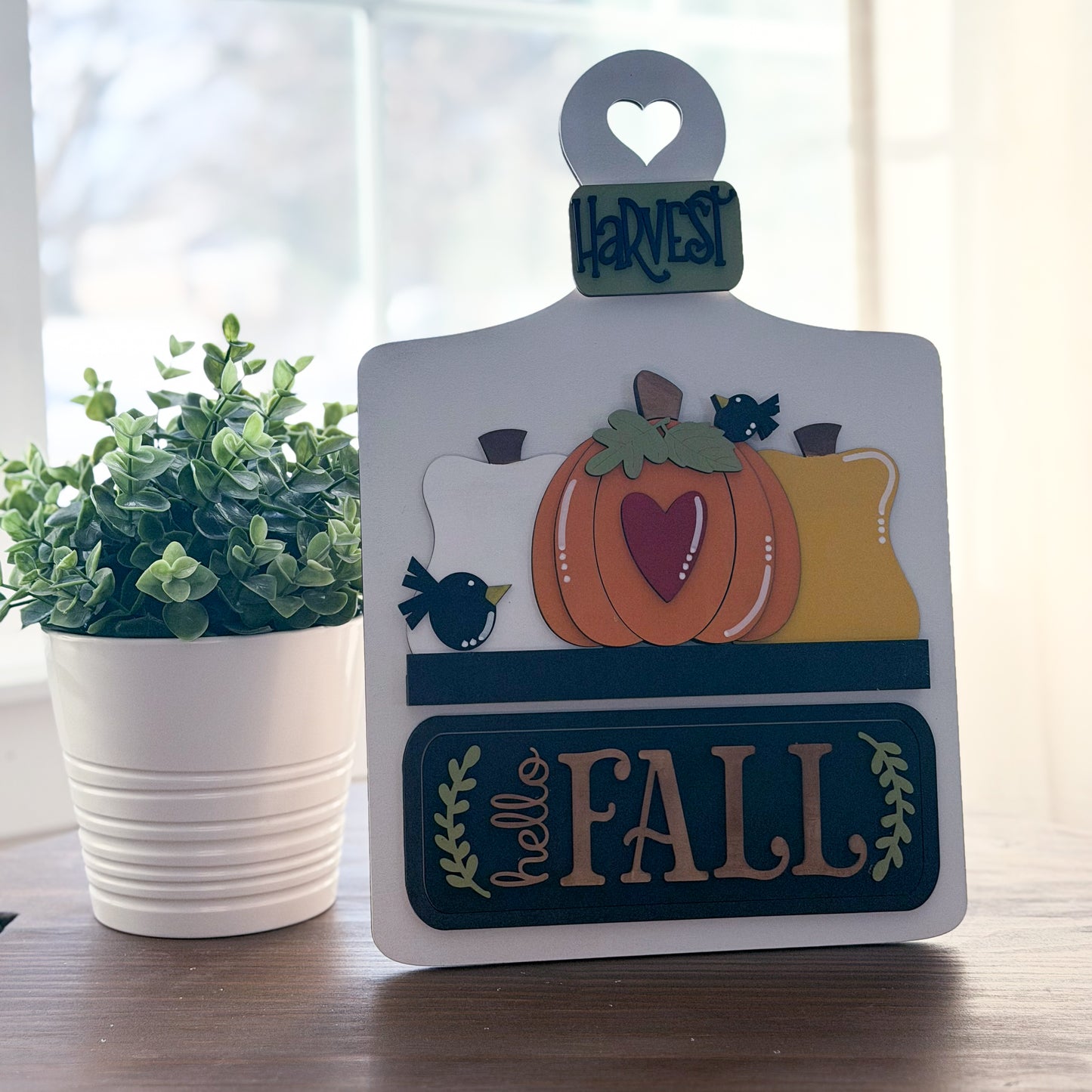 Decorative sign with fall-themed design on a table next to a plant