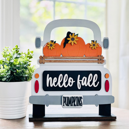 A decorative wood truck with 'hello fall' and 'pumpkin' text, carrying pumpkins on its bed, placed on a table.