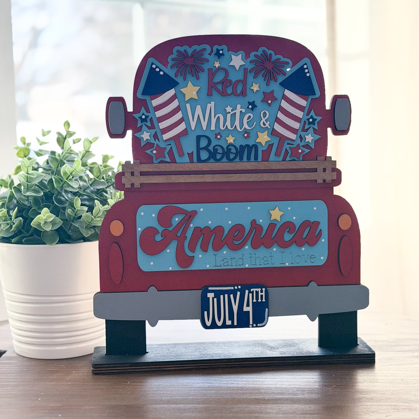 Decorative sign with 'Red White & Boom America Land that I Love' text on a wooden stand.