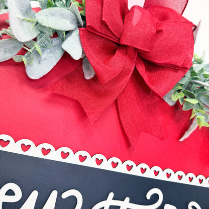 Door sign with a large red bow and greenery, featuring a decorative black and white label.