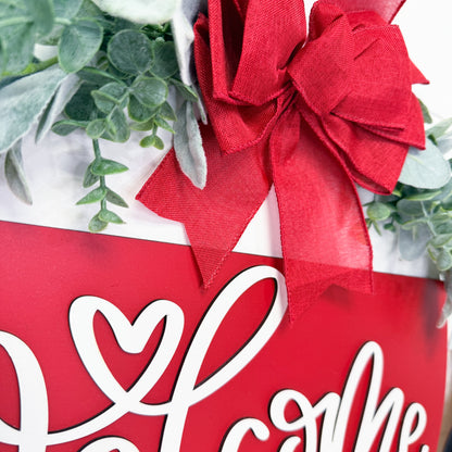 Red bow on a door hanger with white text, surrounded by greenery