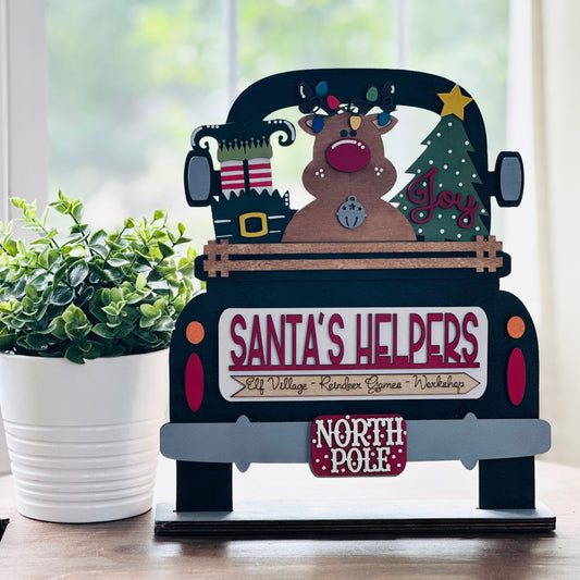 Decorative Christmas truck with reindeer and 'Santa's Helpers' sign on a table.