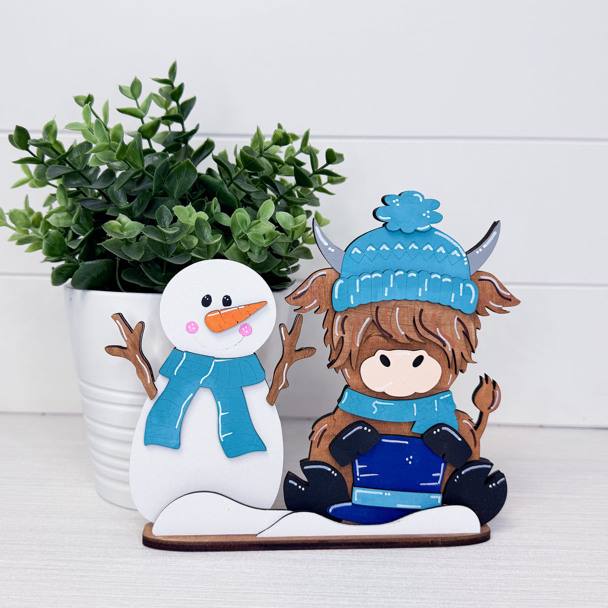 Hand-painted wooden decorative figures of a snowman and a shaggy cartoon bull in winter clothing, displayed on a white surface next to a potted green plant.